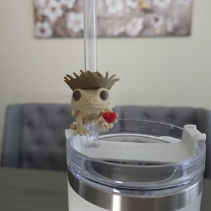 May include: A white Stanley tumbler with a silver rim and a clear straw. A small, tan gecko figurine wearing a brown hat and holding a red heart sits on the straw. The background includes a painting and a gray chair.
