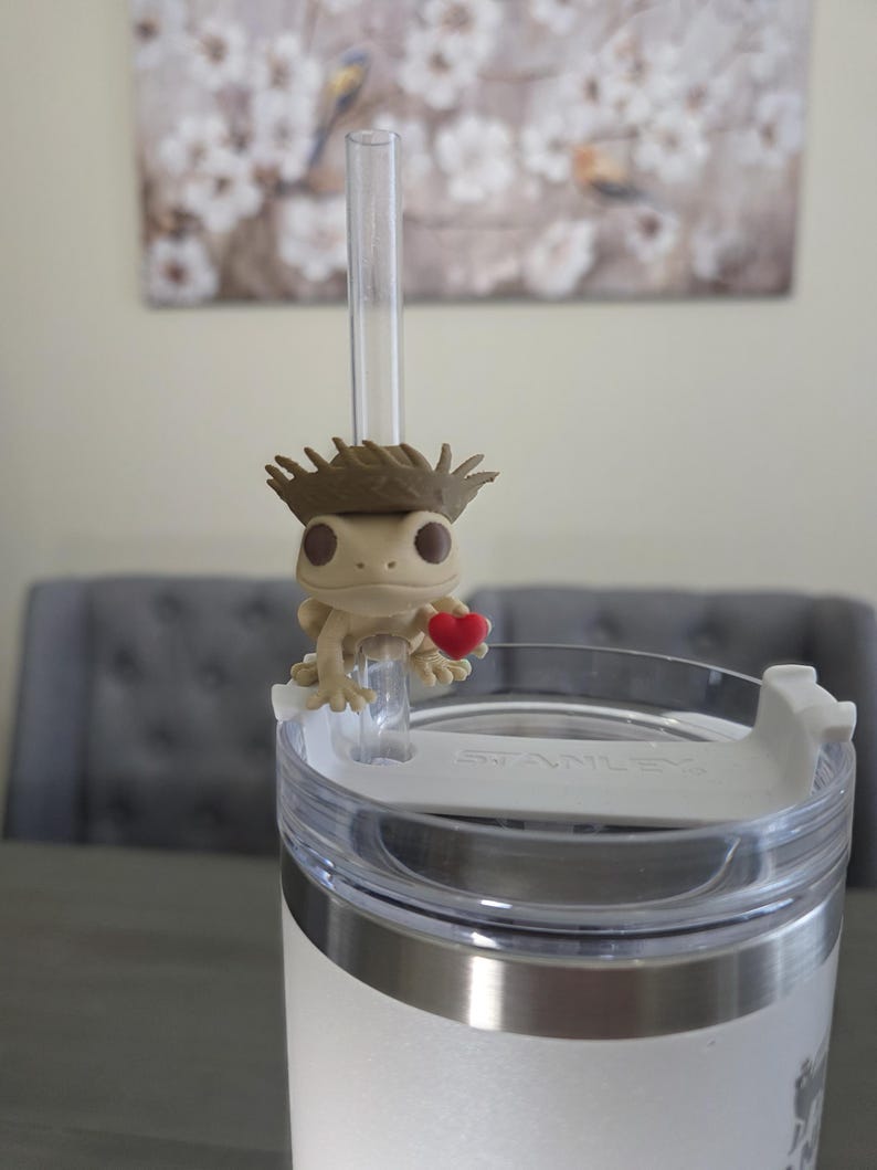 May include: A white Stanley tumbler with a silver rim and a clear straw. A small, tan gecko figurine wearing a brown hat and holding a red heart sits on the straw. The background includes a painting and a gray chair.