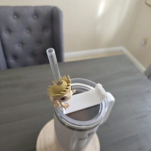 May include: A white and pink insulated tumbler with a clear straw and a decorative figure attached. The tumbler sits on a wooden coaster. The figure is a light brown color with a spiky head.
