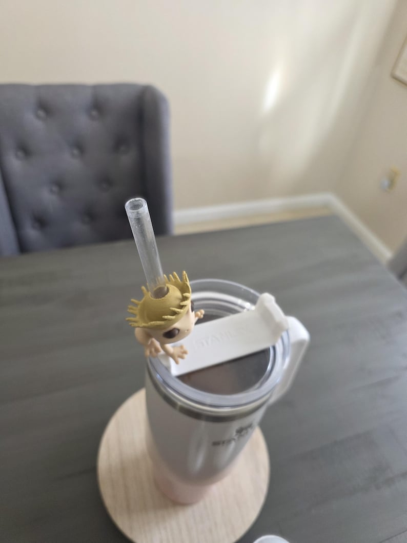 May include: A white and pink insulated tumbler with a clear straw and a decorative figure attached. The tumbler sits on a wooden coaster. The figure is a light brown color with a spiky head.