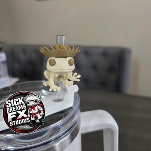 May include: A white tumbler with a clear lid and a white handle. A small, off-white frog figurine wearing a brown hat sits on the lid. The tumbler has a logo that says "SICK DREAMS FX STUDIOS".