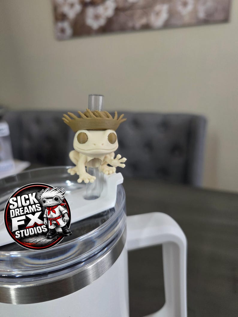 May include: A white tumbler with a clear lid and a white handle. A small, off-white frog figurine wearing a brown hat sits on the lid. The tumbler has a logo that says "SICK DREAMS FX STUDIOS".