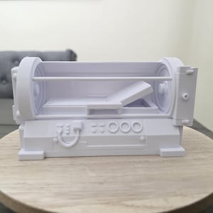 May include: A white, 3D-printed model of a futuristic device, possibly a medical scanner or a piece of science fiction equipment. The model has a rectangular base with circular components on each end and a central compartment. The device is displayed on a wooden surface.
