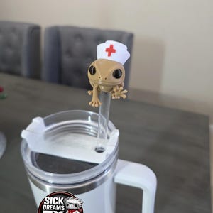 May include: A white travel mug with a handle and a clear lid. A small, tan gecko figurine wearing a nurse's hat with a red cross is attached to a clear straw. The mug has a sticker that says "SICK DREAMS FX STUDIOS."