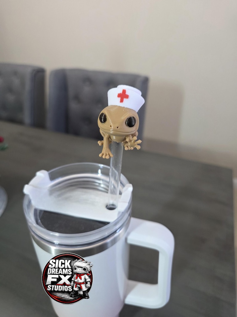 May include: A white travel mug with a handle and a clear lid. A small, tan gecko figurine wearing a nurse's hat with a red cross is attached to a clear straw. The mug has a sticker that says "SICK DREAMS FX STUDIOS."