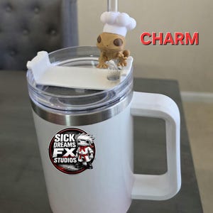 May include: White travel mug with a handle and a clear lid. A small figurine of a lizard wearing a chef's hat sits on top. The mug has a logo that says "SICK DREAMS FX STUDIOS". The word "CHARM" is written in red.