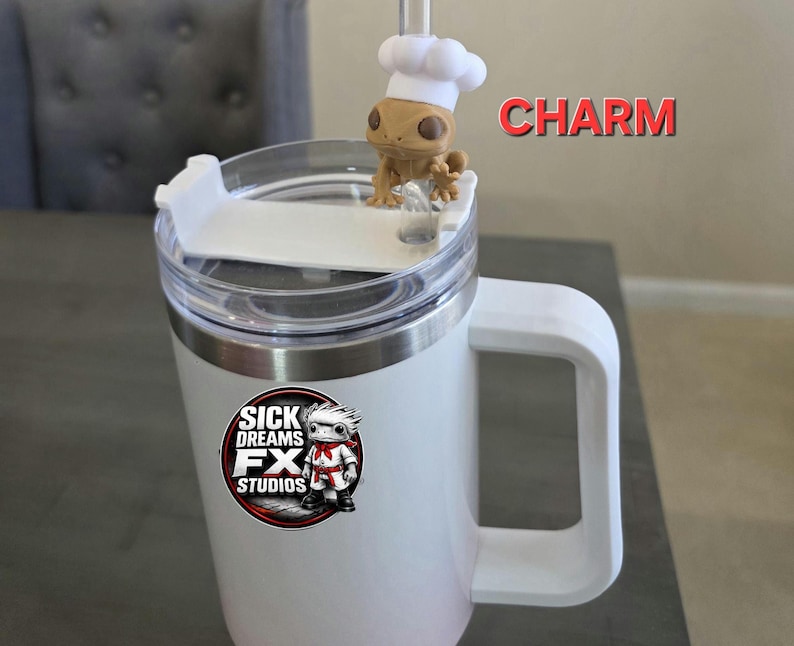 May include: White travel mug with a handle and a clear lid. A small figurine of a lizard wearing a chef's hat sits on top. The mug has a logo that says "SICK DREAMS FX STUDIOS". The word "CHARM" is written in red.
