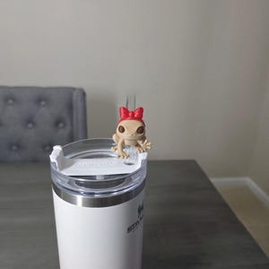 May include: A white and pink ombre Stanley tumbler with a clear lid. A small tan frog figurine with a red bow sits on top of the lid. The tumbler is on a gray wooden table.