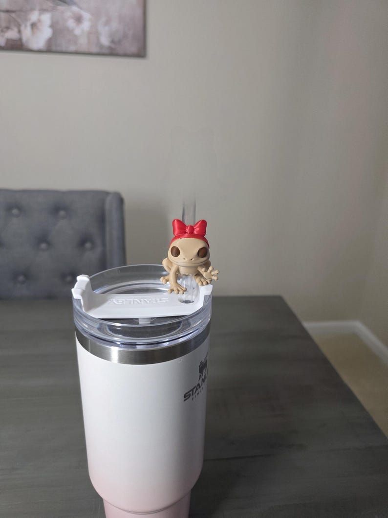 May include: A white and pink ombre Stanley tumbler with a clear lid. A small tan frog figurine with a red bow sits on top of the lid. The tumbler is on a gray wooden table.