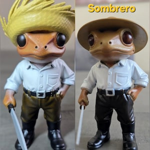 May include: Two frog figurines, one with a yellow straw hat and the word "Pava" above it, and the other with a brown sombrero and the word "Sombrero" above it. Both frogs wear white shirts, dark pants, black boots, and hold a silver tool.
