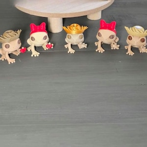 May include: A collection of small, tan-colored frog figurines, each wearing a different hat. The hats include a chef's hat, cowboy hats, a nurse's cap, and others. Some frogs hold small red hearts.