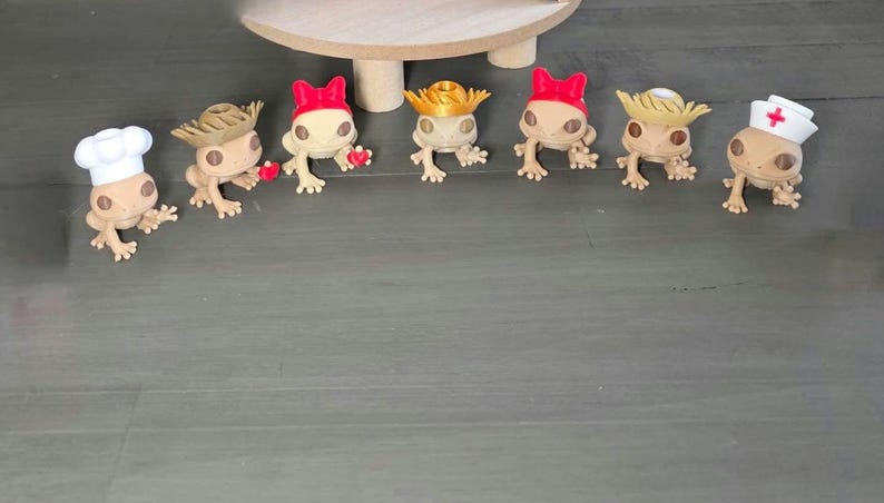 May include: A collection of small, tan figurines of frogs, each wearing a different hat. One frog wears a chef's hat, others wear cowboy hats, a nurse's hat, and other decorative hats. The figurines are arranged on a gray surface.