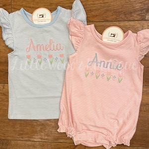 May include: Two striped baby onesies with embroidered names and flowers. The onesie on the left is light blue with the name Amelia and the onesie on the right is pink with the name Annie.