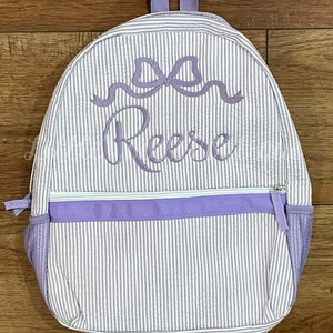 May include: A purple and white striped backpack with a purple bow and the name "Reese" embroidered on the front.