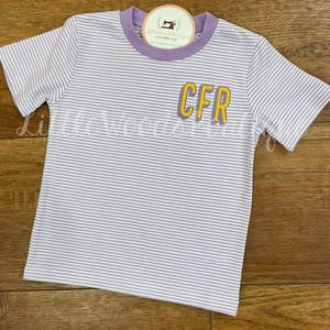 May include: A white and purple striped t-shirt with a purple trim around the neck and a yellow embroidered "CFR" on the front.
