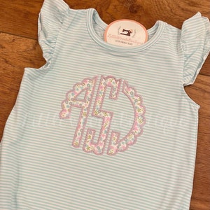 May include: A light blue and white striped baby onesie with a pink and green floral monogram. The onesie has ruffled sleeves and the text "Little Threads Boutique Custom Embroidery & More" is on a white tag.