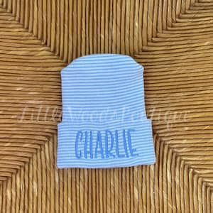 May include: A light blue and white striped newborn beanie with the name "CHARLIE" embroidered in light blue. The hat is made of a soft, ribbed knit material. The background is a woven, light brown surface.