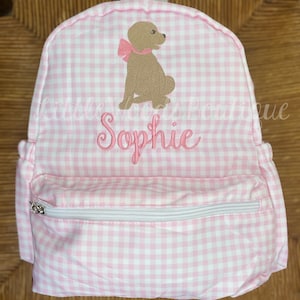 May include: Pink and white gingham fabric backpack with a brown dog embroidered on the front. The backpack has a zippered pocket and the name "Sophie" is embroidered below the dog.