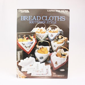 Peut inclure: Une brochure vintage de Leisure Arts intitulée "Bread Cloths Southern Style" par Edi Sweet. La couverture présente une illustration colorée de différents paniers à pain avec des tissus brodés, dont un panier avec un coq et un panier avec un chien.