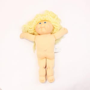 May include: A vintage Cabbage Patch Kid doll with a soft, peach-coloured body and a round face. The doll has blue eyes, a small nose, and a slight smile. Its yellow yarn hair is styled in a messy, fluffy way. The doll is lying on a white surface.