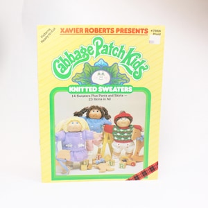 May include: A book cover featuring a yellow background with the text "Xavier Roberts Presents Cabbage Patch Kids Knitted Sweaters" and an illustration of three Cabbage Patch Kids dolls wearing knitted sweaters. The book cover also includes the text "#7866 Plaid".
