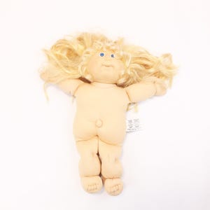 May include: A Cabbage Patch Kid doll with blonde hair and blue eyes. The doll is naked and has a belly button.