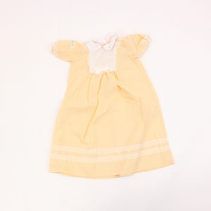May include: A pale yellow baby dress with white lace trim around the neckline and hem. The dress has short sleeves and a white lace collar.