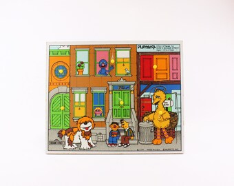 May include: A vintage Playskool Sesame Street puzzle featuring Big Bird, Cookie Monster, and Elmo. The puzzle has 12 pieces and is designed for ages 1 to 4 years. The puzzle depicts a colorful cityscape with a Sesame Street sign.