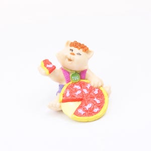 May include: A small, white figurine of a cat wearing a pink and purple shirt and a green necklace with the word "TUG" on it. The cat is holding a slice of pizza with pink fish on it.
