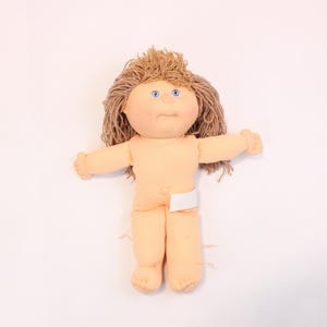 May include: A Cabbage Patch Kid doll with blonde hair and blue eyes. The doll is wearing a white nappy and is lying on its back.
