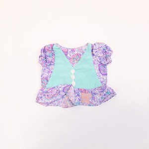 May include: A light blue and purple patterned girls' top with a ruffled hem and a light blue vest with white flower accents.