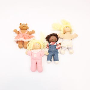 Peut inclure: Quatre poupées Cabbage Patch Kids vintage. Une poupée est un chat brun portant une robe rose. Deux poupées ont les cheveux blonds, l'une en rose et l'autre en short blanc. La quatrième poupée a les cheveux bruns et une salopette bleue.