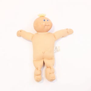 Peut inclure: Une poupée Cabbage Patch Kid avec des cheveux blonds et des yeux bleus. La poupée porte une tenue de couleur chair et a une fleur jaune sur la tête.
