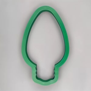 May include: A green plastic cookie cutter in the shape of a Christmas light bulb. The cutter has a detailed base and a smooth, rounded top, perfect for holiday baking. The cutter is set against a plain white background.
