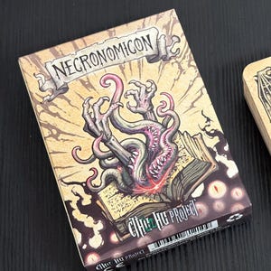 Peut inclure: Boîte rectangulaire avec le mot "NECRONOMICON" en haut. La boîte présente une illustration détaillée d'un monstre avec des tentacules et des griffes sur un livre ouvert. Le texte "Cthulhu Project" est imprimé en bas.