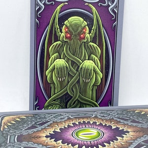 May include: Tarot card featuring a green, tentacled creature with red eyes, titled "The Hierophant". The card has a purple background with ornate gold trim.