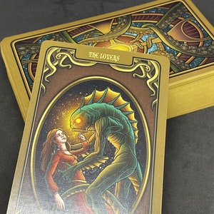 May include: Tarot card depicting The Lovers, featuring a woman in a red dress and a green, fish-like creature with a glowing eye. The card has a gold background with intricate details.
