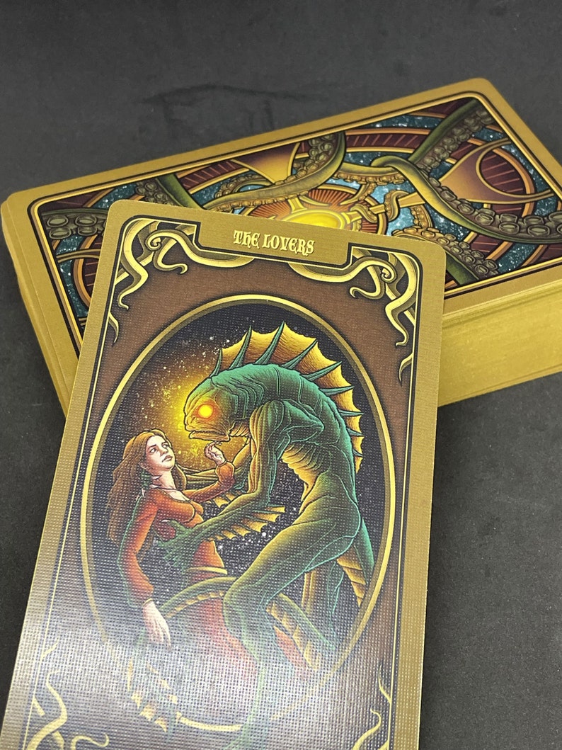 May include: Tarot card depicting The Lovers, featuring a woman in a red dress and a green, fish-like creature with a glowing eye. The card has a gold background with intricate details.