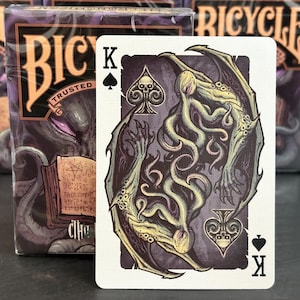 Könnte beinhalten: Eine Bicycle Spielkarte mit einem König Pik Design, das eine Cthulhu-Illustration in Grün, Lila und Creme zeigt. Die Karte zeigt ein schwarzes Pik-Symbol und den Buchstaben "K". Ein Bicycle Kartenspiel ist im Hintergrund zu sehen.