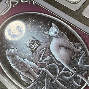May include: A tarot card with a purple background and silver trim. The card features a full moon, a house, and two cats. One cat is sitting on a tentacled creature, while the other cat is looking up at the moon. The card text reads "The Moon".