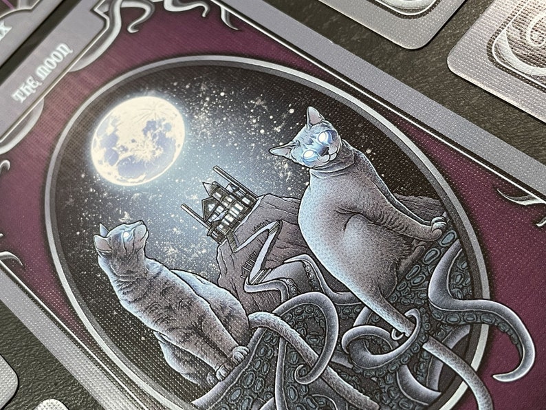 May include: A tarot card with a purple background and silver trim. The card features a full moon, a house, and two cats. One cat is sitting on a tentacled creature, while the other cat is looking up at the moon. The card text reads "The Moon".