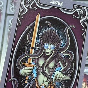 May include: A tarot card with a purple background and a silver border. The card depicts a green-skinned creature with a long, flowing tail and a sword in one hand and a scale in the other. The card is titled "Justice".