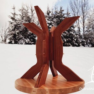 Puede incluir: Una base de mesa de madera con un diseño geométrico único. La base está hecha de madera marrón rojiza y tiene una parte superior circular. La base está hecha de cuatro piezas de madera que se unen de forma que crean una forma de estrella. La base de la mesa está sobre un suelo nevado con árboles en el fondo.