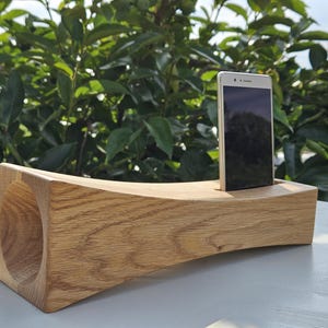 Può includere: Un supporto per telefono in legno con un design curvo e una grande apertura circolare. Uno smartphone è posizionato nel supporto. Il supporto è realizzato in legno chiaro e poggia su una superficie bianca, con fogliame verde sullo sfondo.