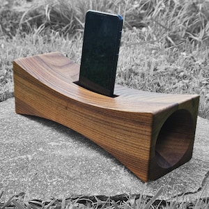 Puede incluir: Soporte de teléfono de madera marrón claro con diseño curvo y ranura para sostener el teléfono. El soporte tiene una abertura circular en un extremo. El teléfono es negro.