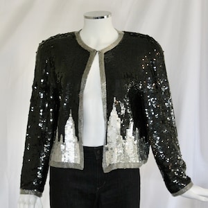 May include: A black and silver sequined jacket with a silver trim. The jacket has a long sleeve and a front closure.