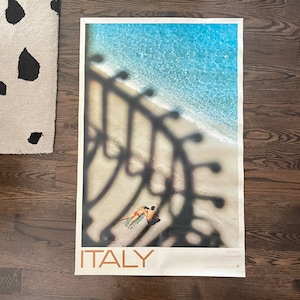 May include: A vintage travel poster for Italy featuring a beach scene with two people sunbathing. The poster has a blue and white color scheme and features the text "ITALY" and "TROPEA (Calabria)".