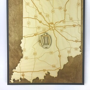 May include: A wooden map of Indiana with a 3D model of Lucas Oil Stadium in the center. The map features highway numbers and the state outline. The background is a dark brown wood.