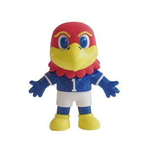 May include: A small figurine of a bird mascot with a red head, yellow beak, and blue eyes. It wears a blue jersey with the number 1 printed in white on the chest, white shorts, and yellow shoes. The figure has outstretched arms and a smiling face.