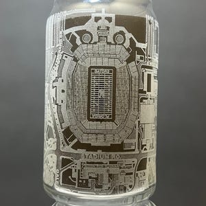 May include: A clear glass can with an etched design of a football stadium. The stadium is labeled "Ben Hill Griffin Stadium" and "Gators".